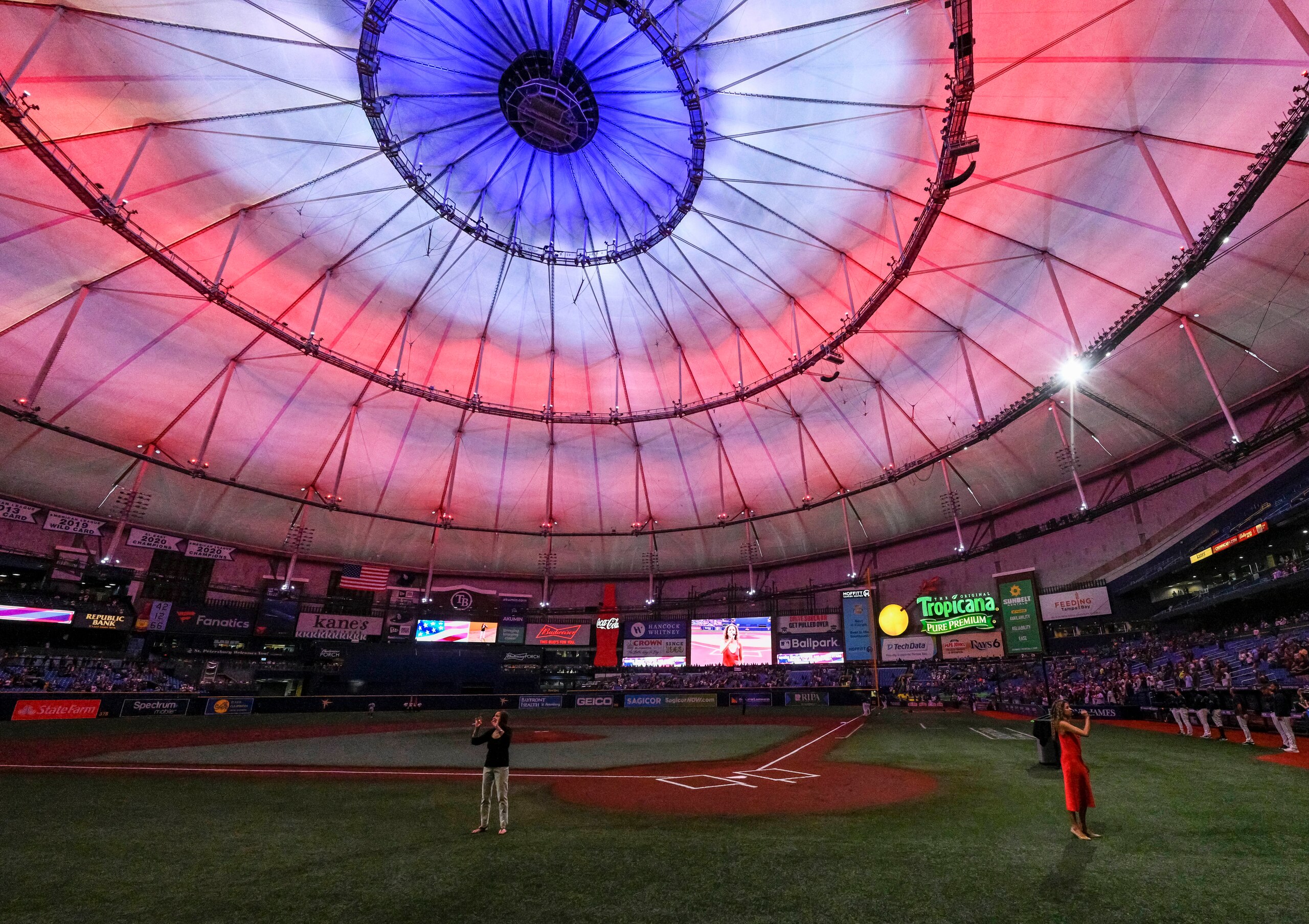 A singer, right, performs the national anthem below red, white and blue lights at Tropicana Field before a baseball game between the Tampa Bay Rays and Miami Marlins Friday, Sept. 24, 2021, in St. Petersburg, Fla. (AP Photo/Steve Nesius)