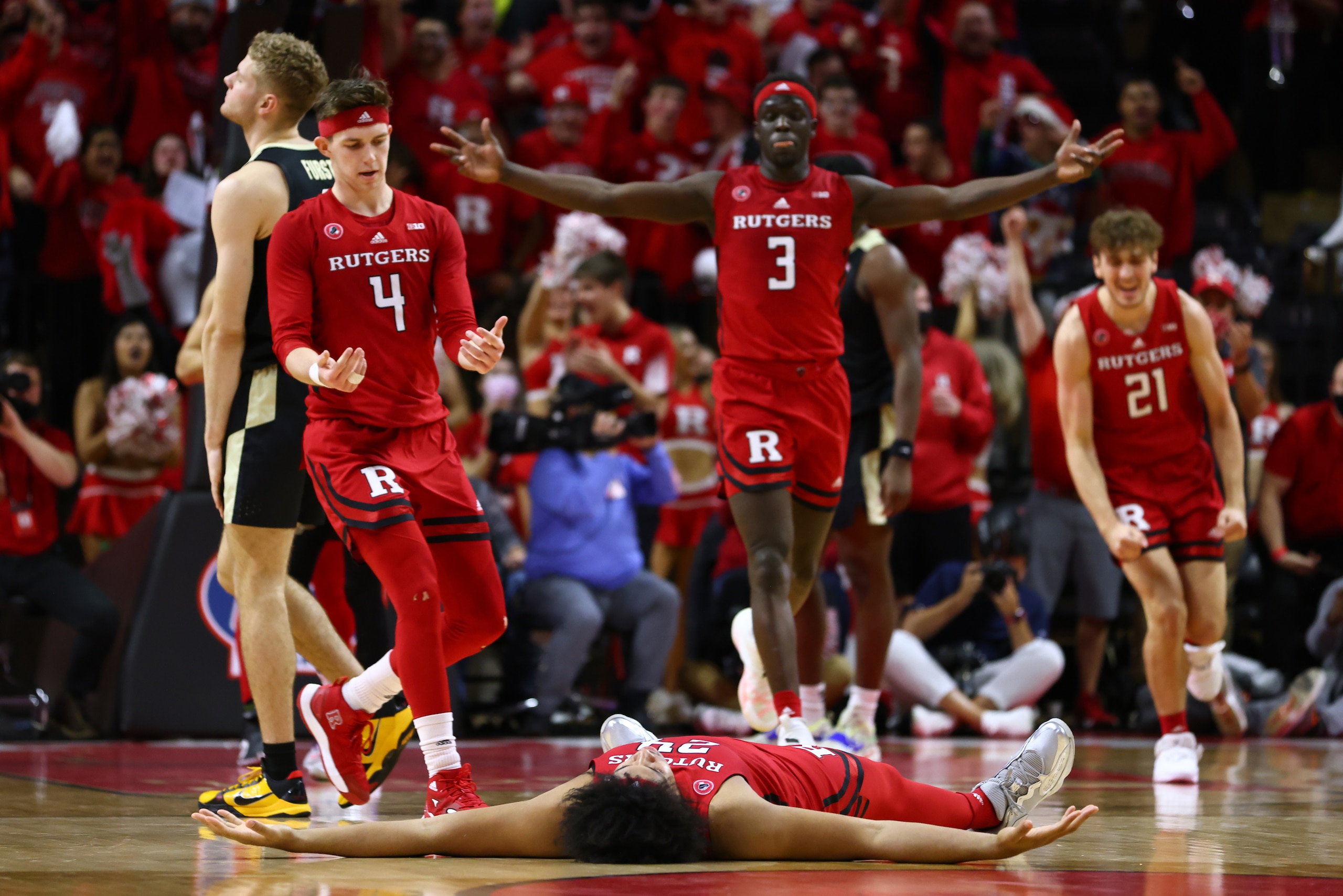 PISCATAWAY, NJ - DECEMBER 09:  Ron Harper Jr. #24 of the Rutgers Scarlet Knights lies on the court after sinking a three-point shot as teammates Paul Mulcahy #4, Mawot Mag #3 and Dean Reiber #21celebrate during the first half of a game at Jersey Mike's Arena on December 9, 2021 in Piscataway, New Jersey. (Photo by Rich Schultz/Getty Images)