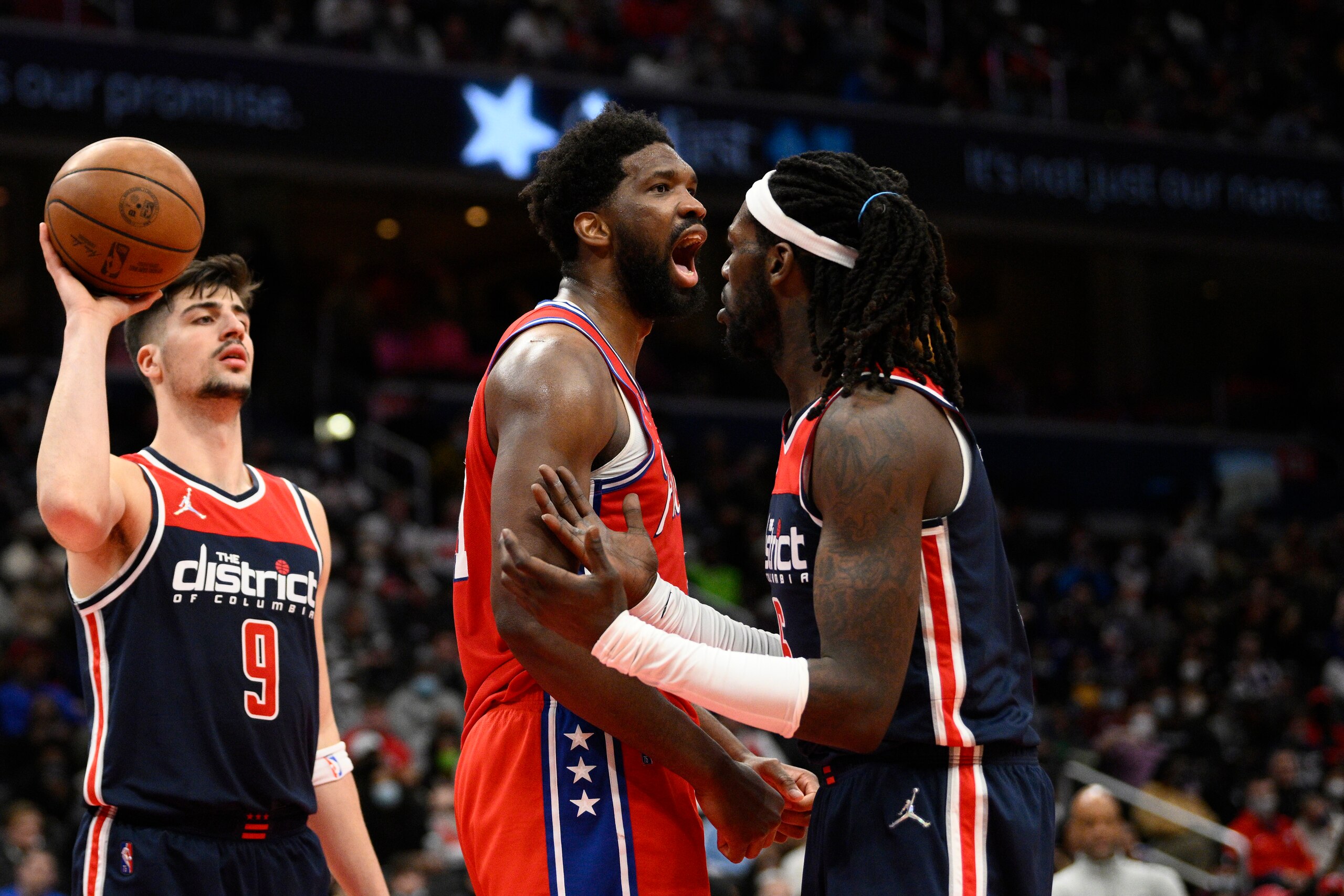 Philadelphia 76ers center Joel Embiid, center, reacts after he was fouled by Washington Wizards center Montrezl Harrell, right, during the second half of an NBA basketball game, Sunday, Dec. 26, 2021, in Washington. Wizards forward Deni Avdija is at left. The 76ers won 117-96. (AP Photo/Nick Wass)