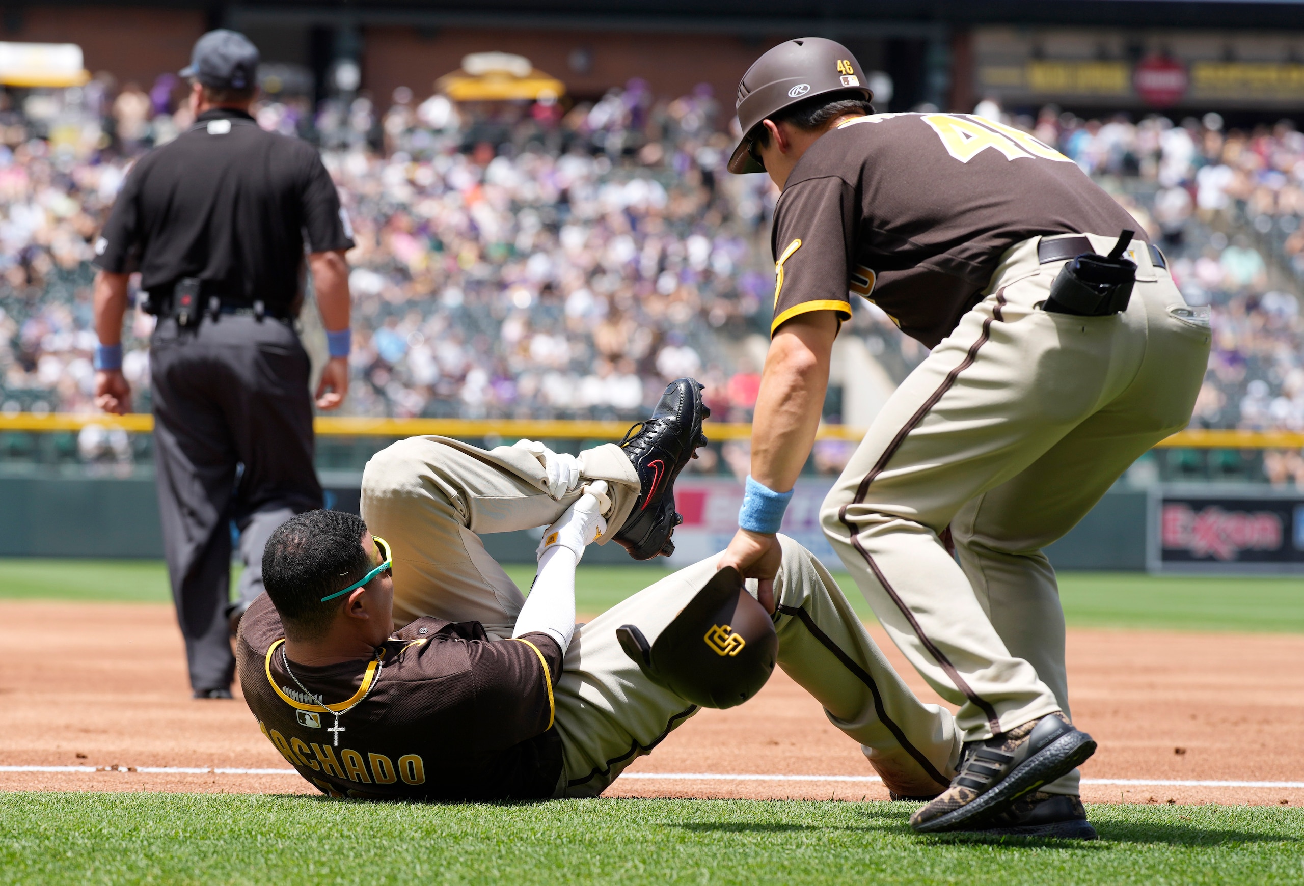 San Diego Padres first base coach David Macias, right, looks to help Manny Machado after Machado was injured while trying to run out an infield hit to Colorado Rockies starting pitcher Antonio Senzatela in the first inning of a baseball game Sunday, June 19, 2022, in Denver. Machado, who was out on the play, had to be helped from the field. (AP Photo/David Zalubowski)