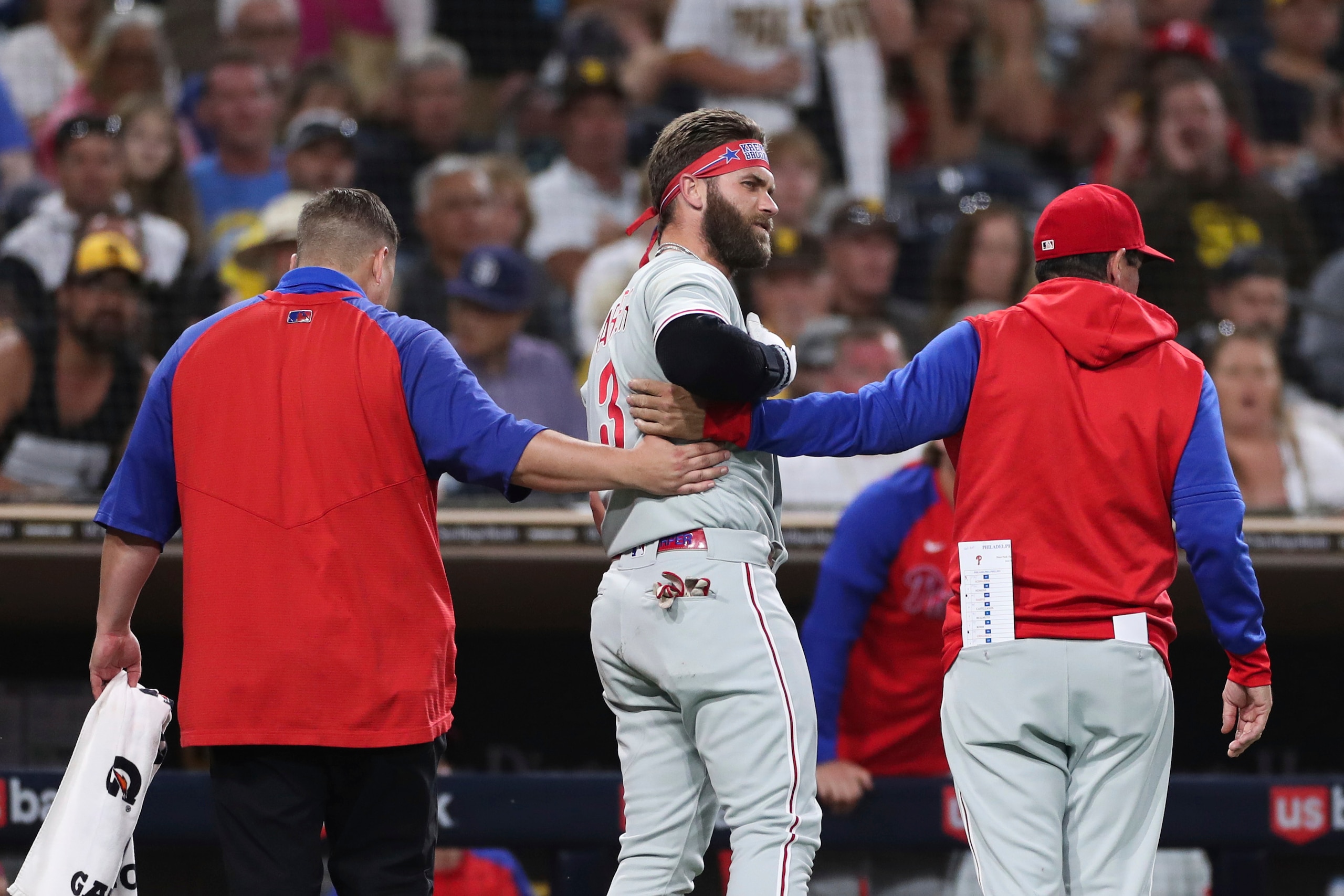Philadelphia Phillies' Bryce Harper, center, reacts after being hit by a pitch as he walks off the field with interim manager Rob Thomson, right, and a trainer, left in the fourth inning of a baseball game against the San Diego Padres, Saturday, June 25, 2022, in San Diego. (AP Photo/Derrick Tuskan)