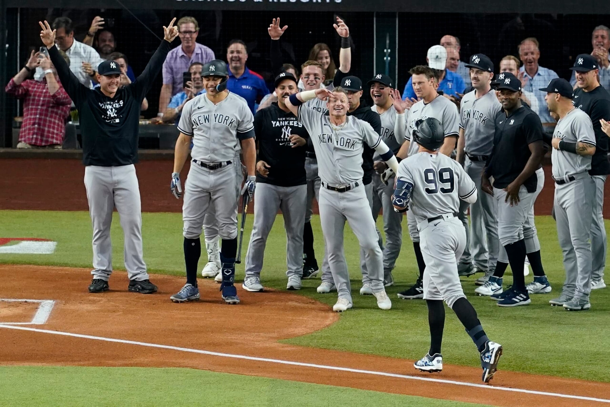 New York Yankees' Aaron Judge (99) approaches home plate as teammates come out to congratulate him after hitting a solo home run, his 62nd of the season, in the first inning of the second baseball game of a doubleheader against the Texas Rangers in Arlington, Texas, Tuesday, Oct. 4, 2022. With the home run, Judge set the AL record for home runs in a season, passing Roger Maris. Umpire Randy Rosenberg and Rangers catcher Sam Huff also stand by watching the play. (AP Photo/Tony Gutierrez)