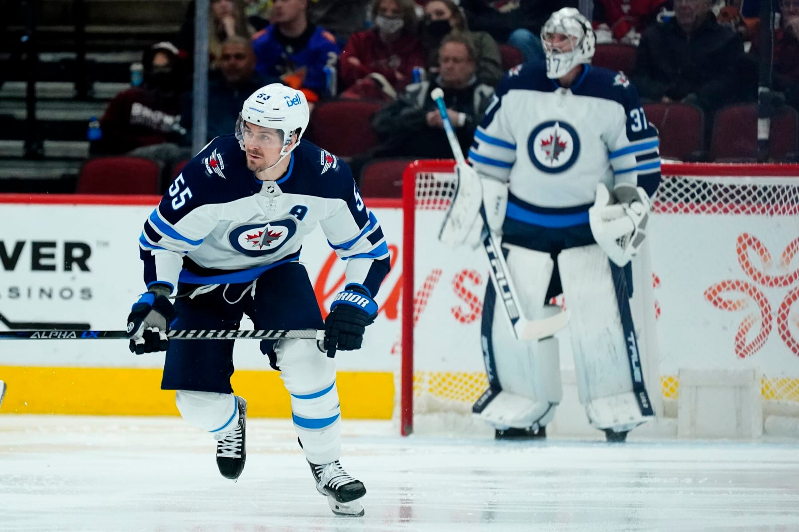 Winnipeg Jets center Mark Scheifele skates up ice as goaltender Connor Hellebuyck looks on.