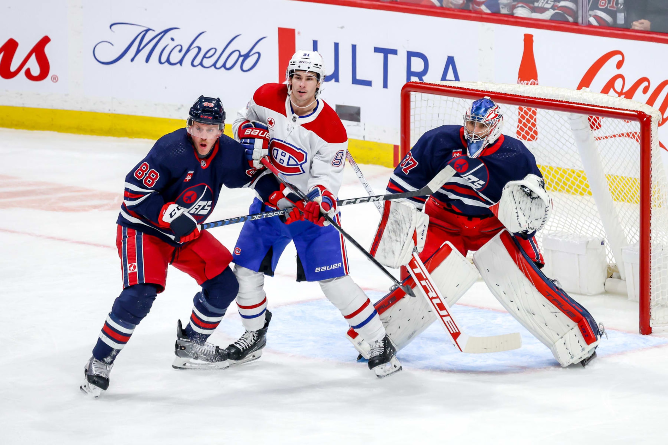 WINNIPEG, CANADA - DECEMBER 18: Sean Monahan #91 of the Montreal Canadiens battles Nate Schmidt #88 and goaltender Connor Hellebuyck #37 of the Winnipeg Jets as they keep an eye on the play during first period action at the Canada Life Centre on December 18, 2023 in Winnipeg, Manitoba, Canada. (Photo by Jonathan Kozub/NHLI via Getty Images)