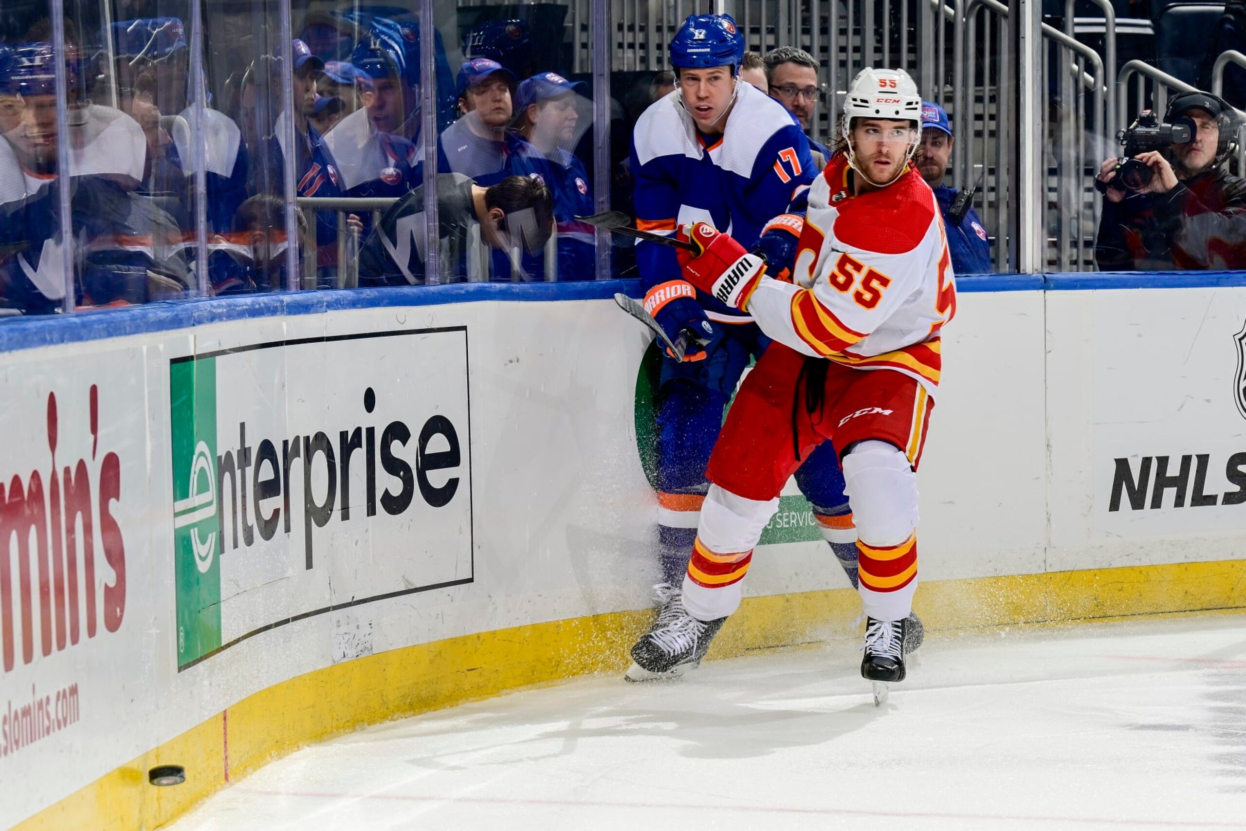 ELMONT, NEW YORK - FEBRUARY 10:  Matt Martin #17 of the New York Islanders and Noah Hanifin #55 of the Calgary Flames pursue the puck during the third period at UBS Arena on February 10, 2024 in Elmont, New York. (Photo by Steven Ryan/NHLI via Getty Images)