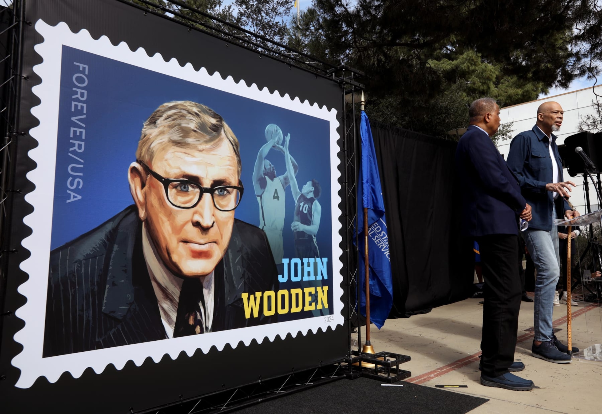 LOS ANGELES,  CA  -  FEBRUARY 22, 2024 - - Lakers great and UCLA alum Kareem Abdul-Jabbar, right, standing next to Basketball Hall of Famer Jamaal Wilkes, makes a presentation at the first-day-of-issue ceremony for a commemorative Forever stamp depicting the late UCLA basketball coach John Wooden held Saturday outside Pauley Pavilion on the UCLA campus in Los Angeles on February 22, 2024. UCLA Chancellor Gene Block, former Bruins' women's gymnastics coach Valorie Kondos Field, Christy Impelman, a granddaughter of Wooden, and Derek Kan, a member of the U.S. Postal Service Board of Governors made presentations to the late UCLA coach. (Genaro Molina/Los Angeles Times via Getty Images)