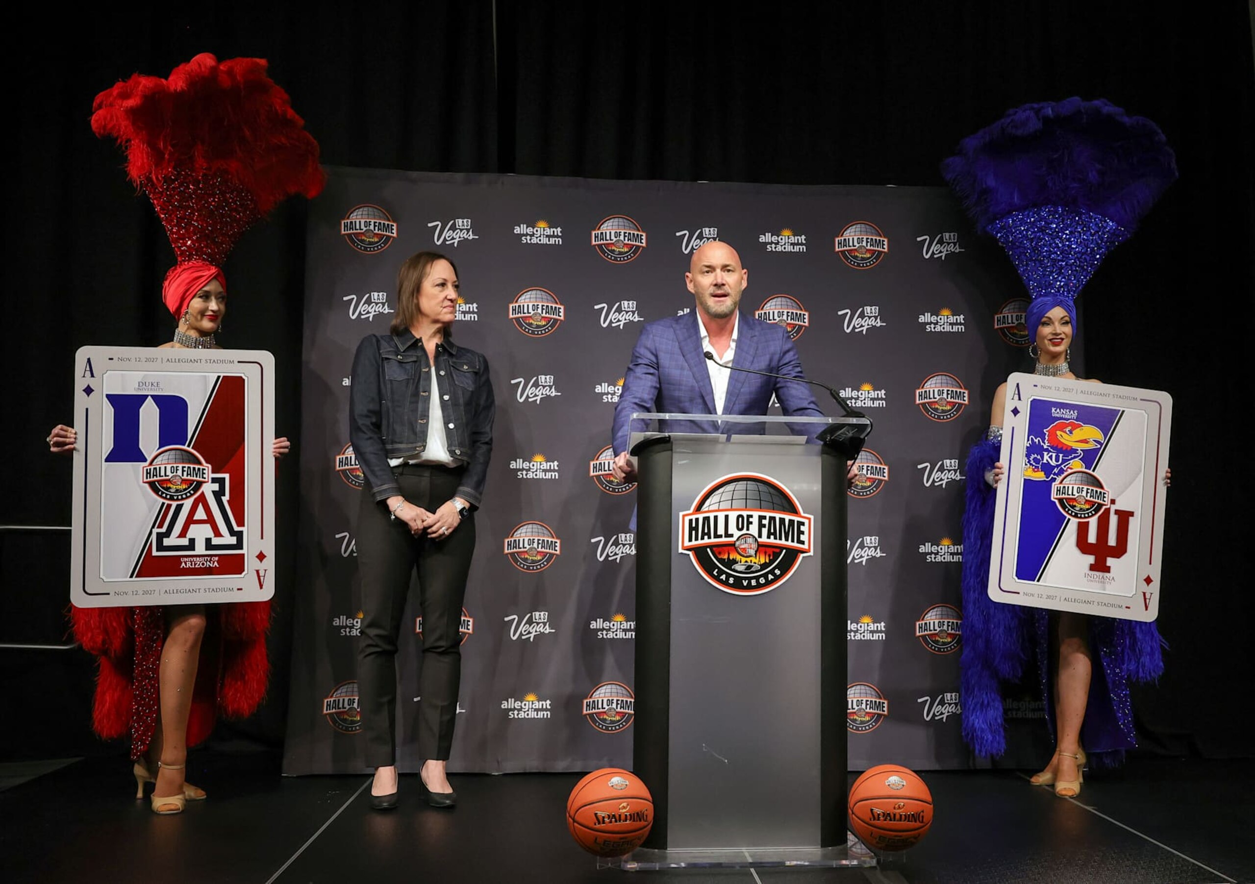 LAS VEGAS, NEVADA - OCTOBER 27: Position Sports President Melissa Meacham-Grossman (2nd L) looks on as Naismith Basketball Hall of Fame President and CEO John L. Doleva, flanked by showgirls holding oversized playing cards with team logos, speaks during a news conference to announce the 2027 Hall of Fame Series Las Vegas at Allegiant Stadium on October 27, 2024 in Las Vegas, Nevada. The doubleheader will mark the first-ever basketball game played at Allegiant Stadium and will feature matchups between Arizona and Duke and Indiana and Kansas on November 12, 2027. (Photo by Ethan Miller/Getty Images)