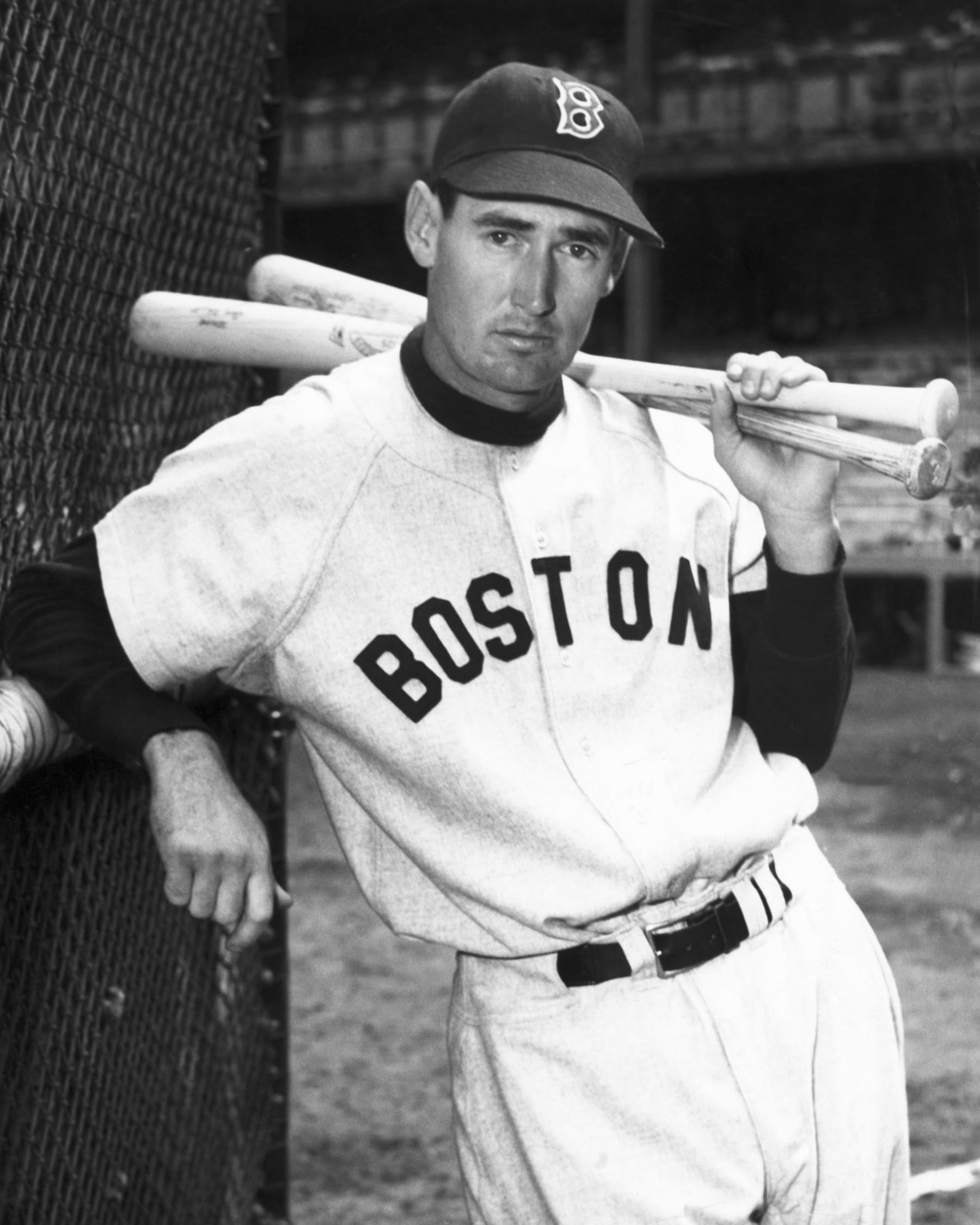 Ted Williams, Boston Red Sox slugger, leans against a fence with two bats over his shoulder.