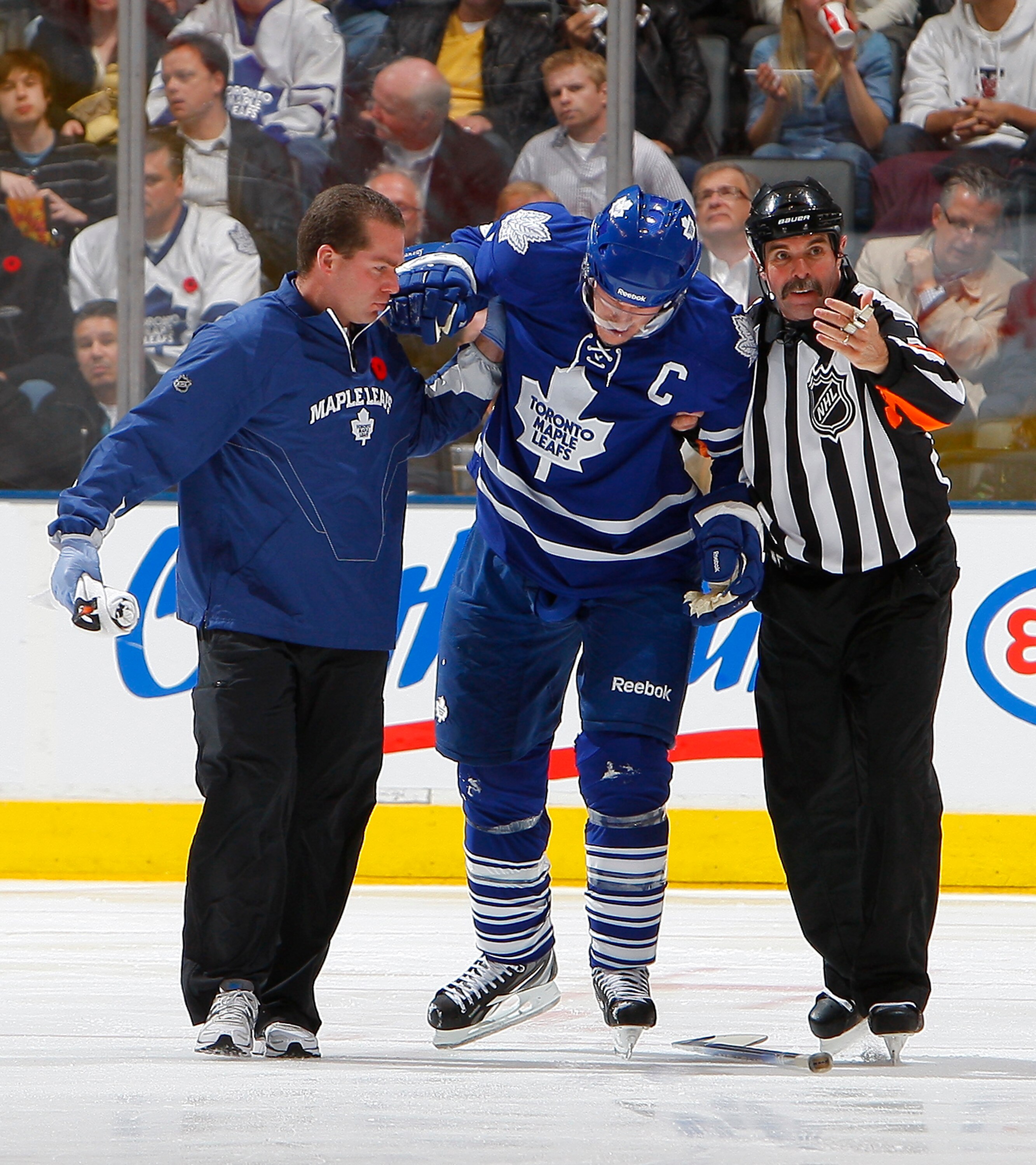 TORONTO - NOVEMBER 2:  Dion Phaneuf #3 of the Toronto Maple Leafs is taken off the ice by head athletic therapist  Andy Playter and referee Bill McCreary during NHL action against the Ottawa Senators at the Air Canada Centre November 2, 2010 in Toronto, O