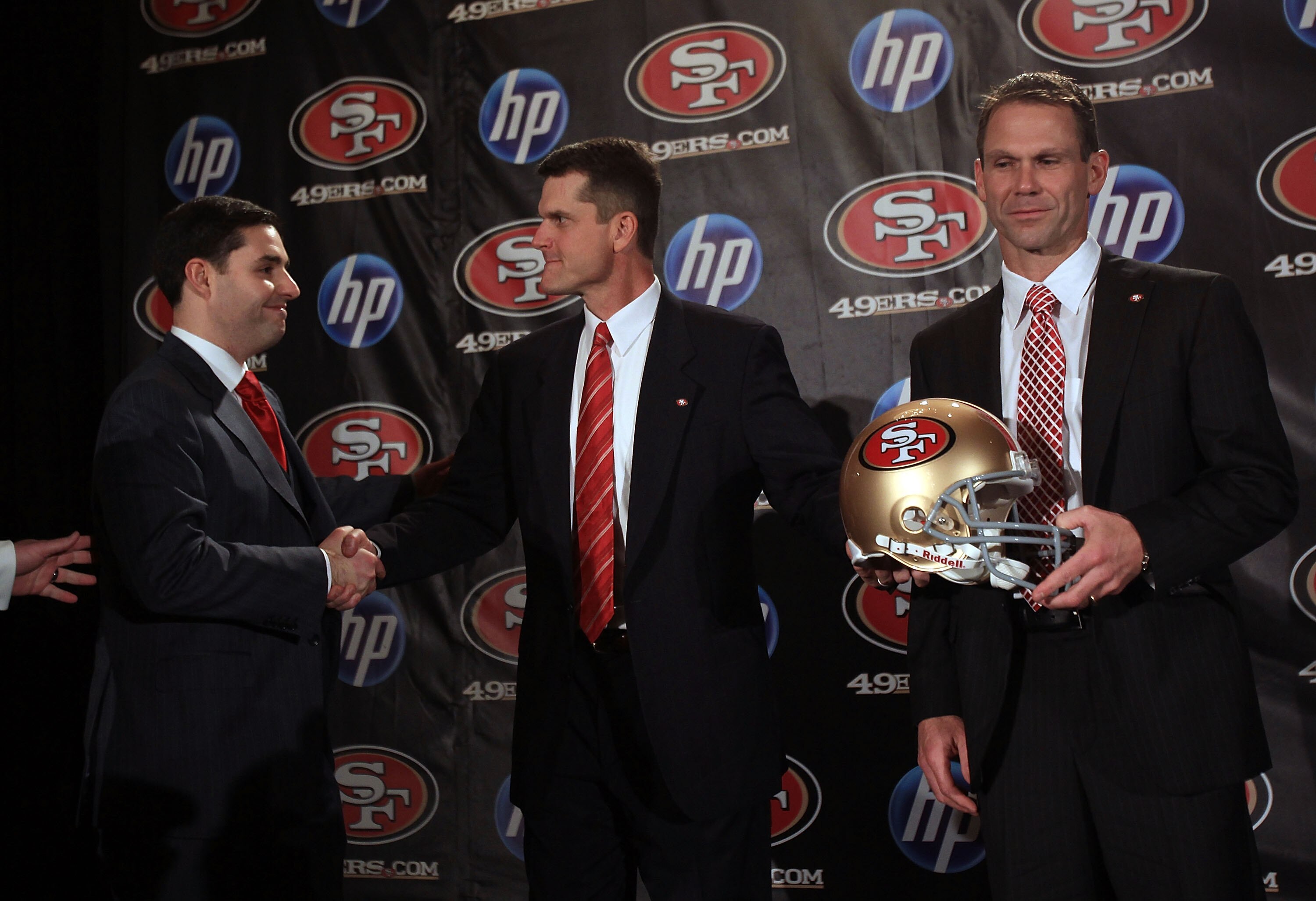 SAN FRANCISCO, CA - JANUARY 07:  Jim Harbaugh shakes hands with President and CEO of the San Francisco 49ers, Jed York, while general manager Trent Baalk holds a 49ers helmet following a press conference where Harbaugh was introduced as the new San Franci