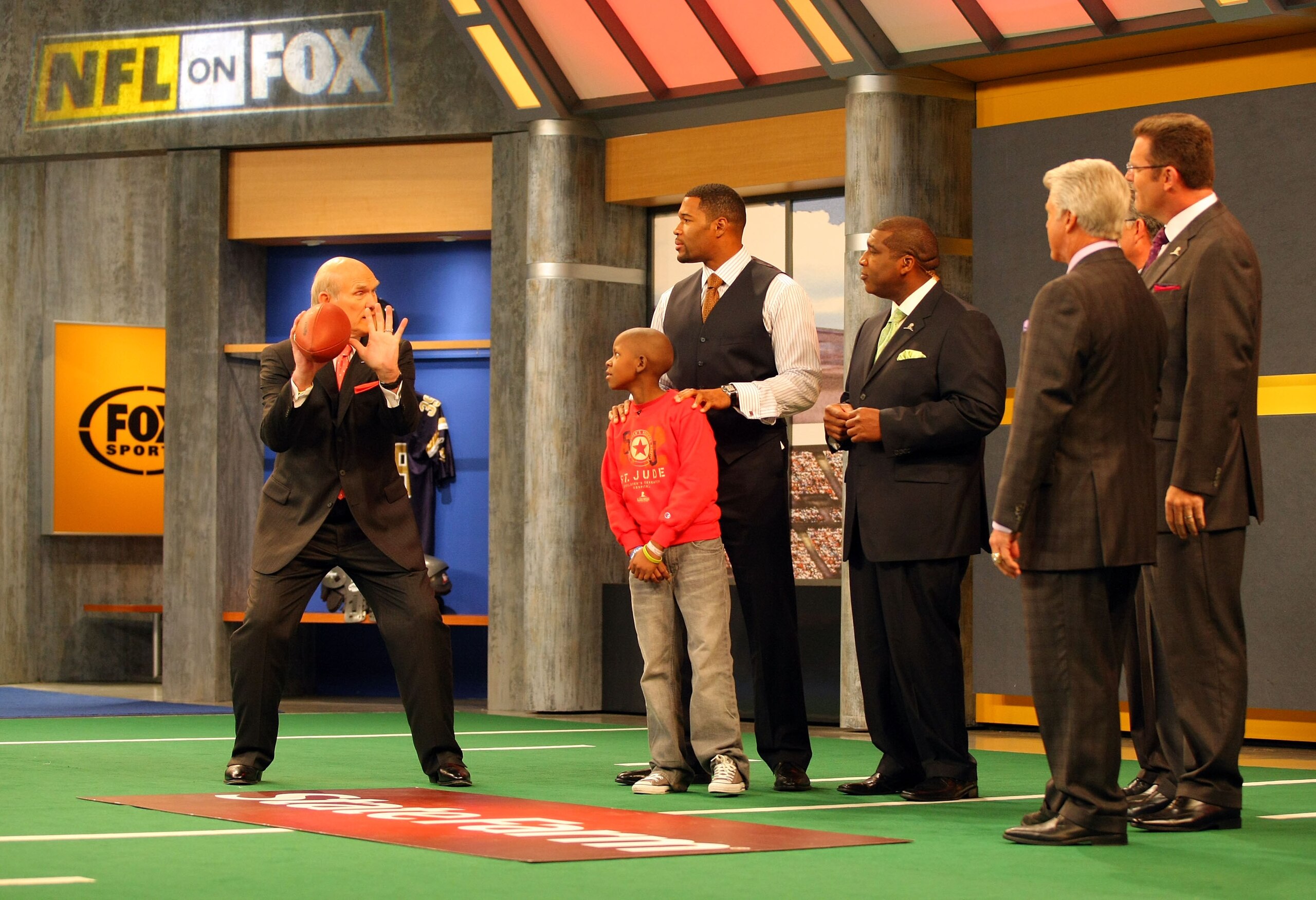 LOS ANGELES, CA - DECEMBER 12:  (L-R) FOX NFL Sunday host Terry Bradshaw demonstrates a move to St. Jude Children's Research Hospital patient Markell, analyst Michael Strahan, host Curt Menefee, analysts Jimmy Johnson and Howie Long and former NFL referee
