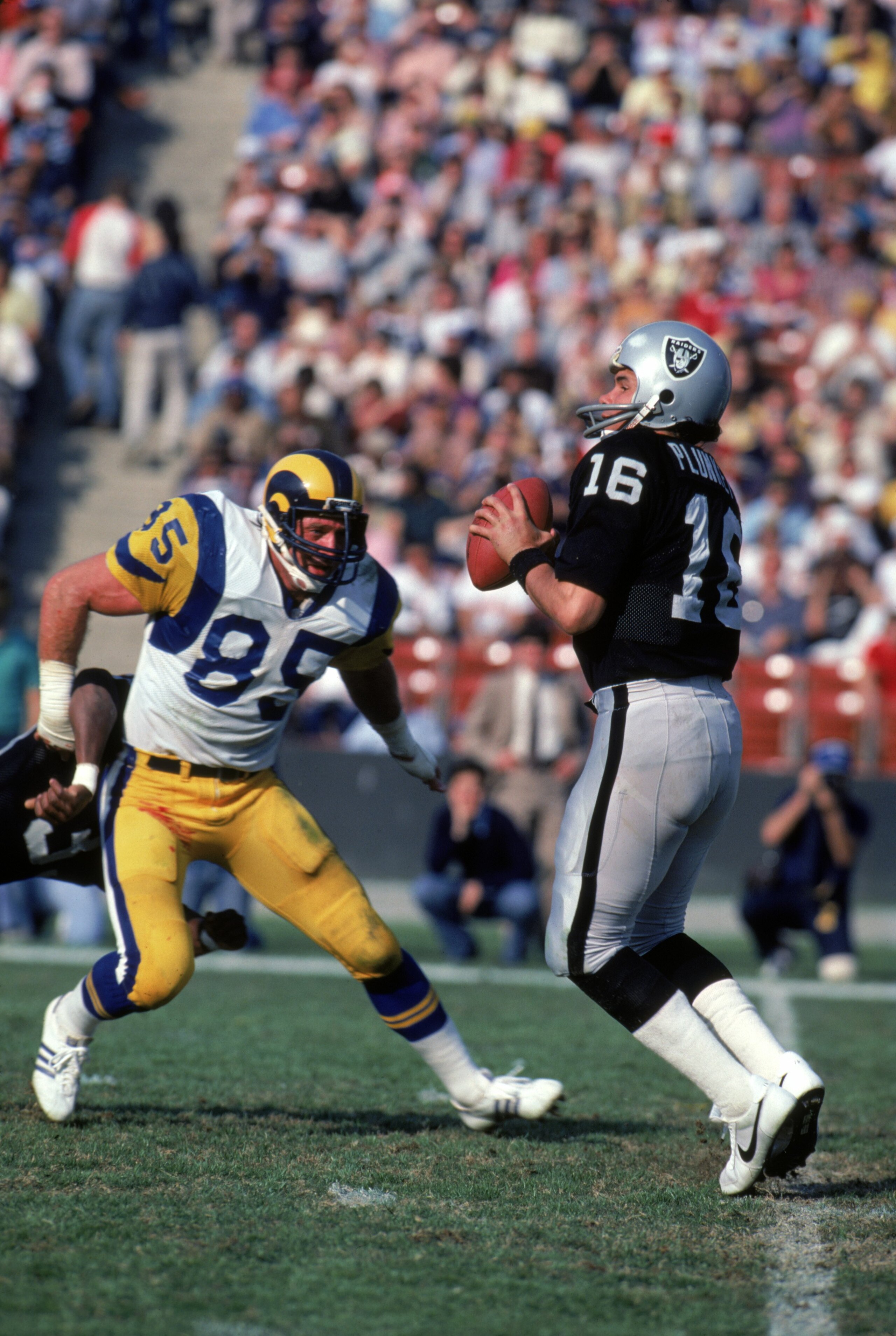 LOS ANGELES - DECEMBER 18:  Quarterback Jim Plunkett #16 of the Los Angeles Raiders looks down field for a receivers as he gets rushed by Los Angeles Rams defensive end Jack Youngblood #85 during a game at Los Angeles Memorial Coliseum on December 18, 198