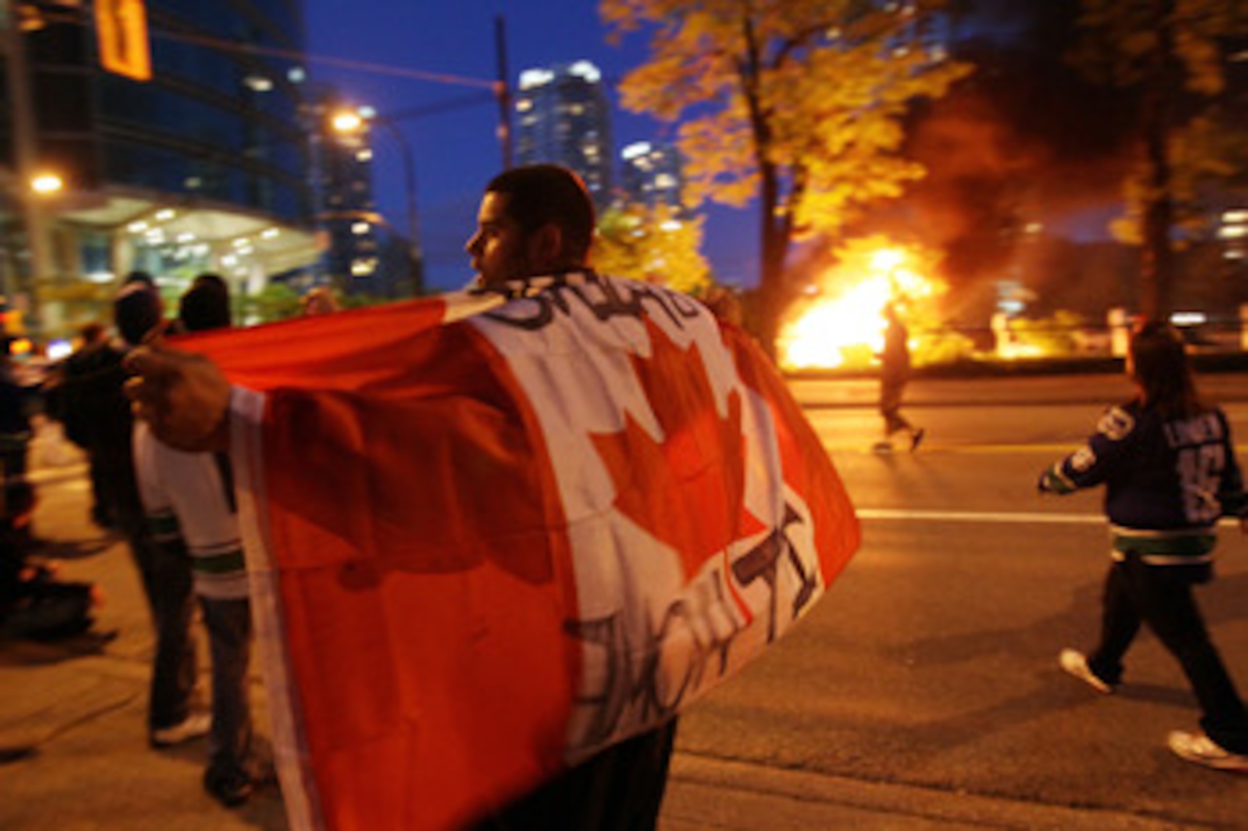 VANCOUVER, BC - JUNE 15:  A person with a Candanian flag walks in front of a burning vehicle on June 15, 2011 in Vancouver, Canada. Vancouver broke out in riots after their hockey team the Vancouver Canucks lost in Game Seven of the Stanley Cup Finals.  (