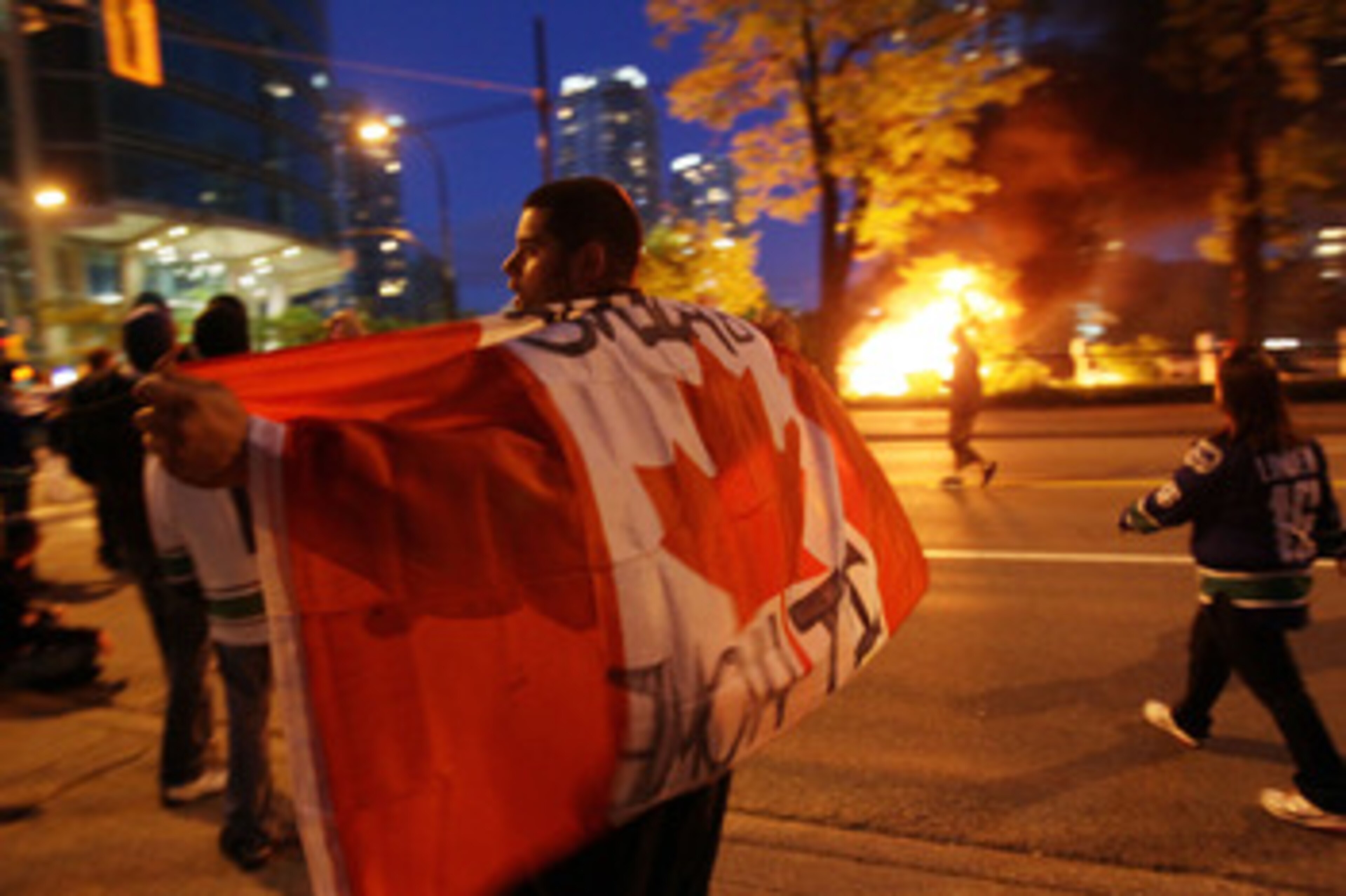 VANCOUVER, BC - JUNE 15:  A person with a Candanian flag walks in front of a burning vehicle on June 15, 2011 in Vancouver, Canada. Vancouver broke out in riots after their hockey team the Vancouver Canucks lost in Game Seven of the Stanley Cup Finals.  (
