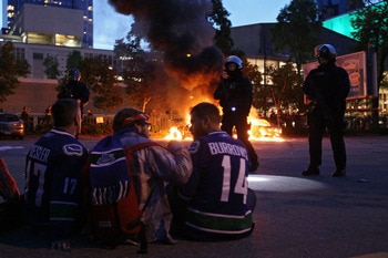 VANCOUVER, BC - JUNE 15:  Riot police stand in front of two burning police cars as people look on June 15, 2011 in Vancouver, Canada. Vancouver broke out in riots after their hockey team the Vancouver Canucks lost in Game Seven of the Stanley Cup Finals. 