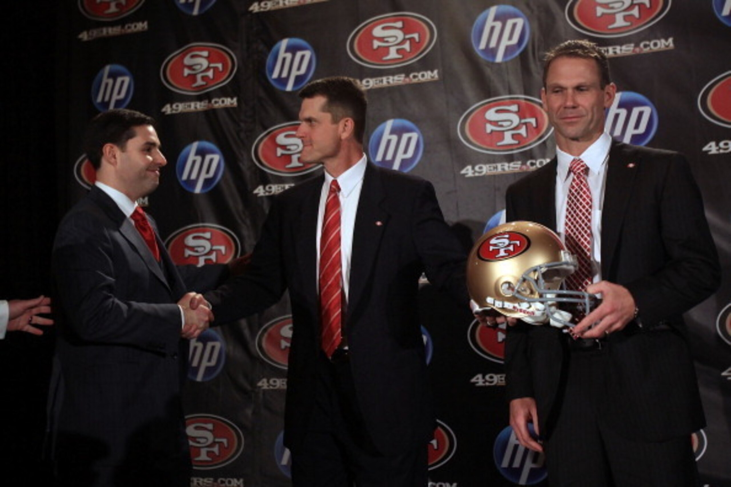 SAN FRANCISCO, CA - JANUARY 07:  Jim Harbaugh shakes hands with President and CEO of the San Francisco 49ers, Jed York, while general manager Trent Baalk holds a 49ers helmet following a press conference where Harbaugh was introduced as the new San Franci