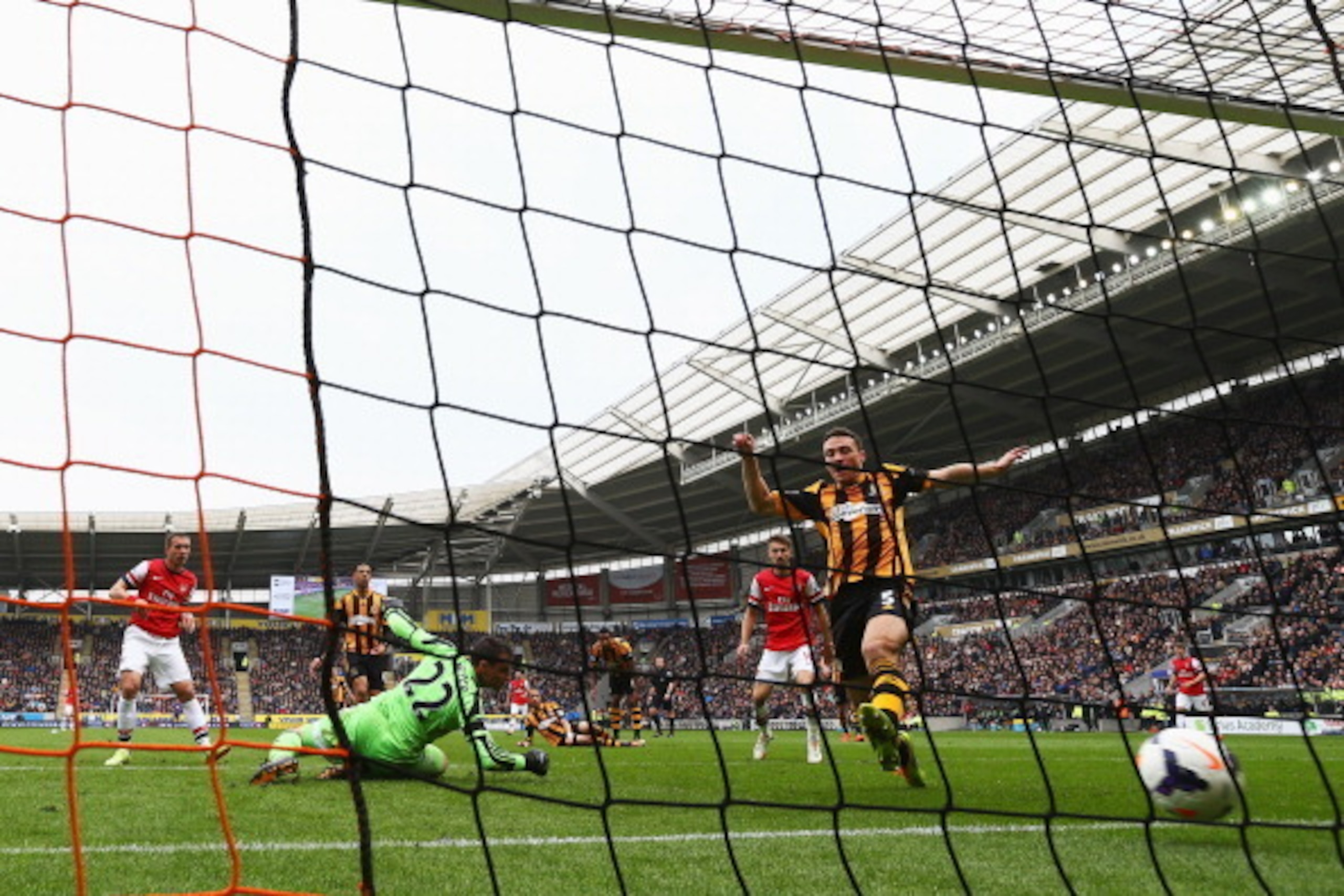 HULL, ENGLAND - APRIL 20:  Lukas Podolski (L) of Arsenal shoots past Steve Harper the Hull City goalkeeper and James Chester (R) of Hull City to score his sides third goal during the Barclays Premier League match between Hull City and Arsenal at KC Stadiu