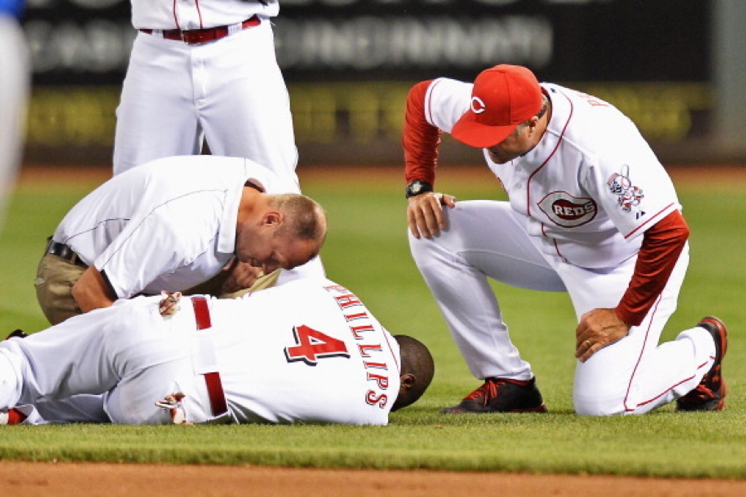 CINCINNATI, OH - JULY 9: Athletic trainer Steve Baumann of the Cincinnati Reds attends to Brandon Phillips #4 of the Cincinnati Reds and Manager Bryan Price #38 of the Cincinnati Reds watches after Phillips injured his thumb diving for a ball in the eight