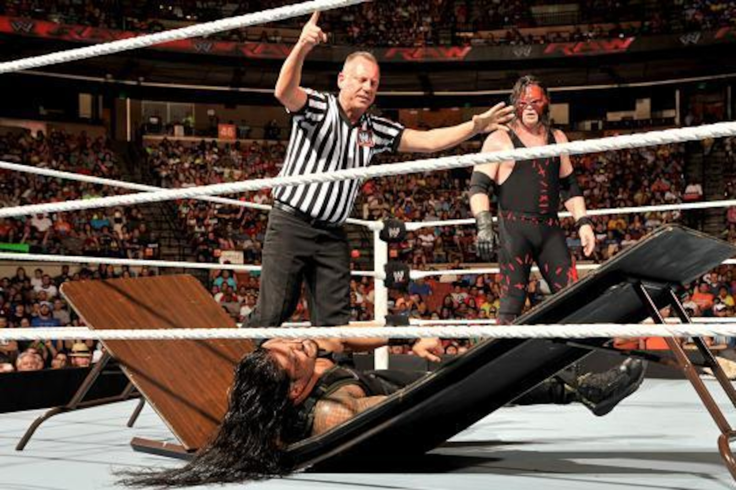 Kane looks on as Roman Reigns tries to get up from a chokeslam.