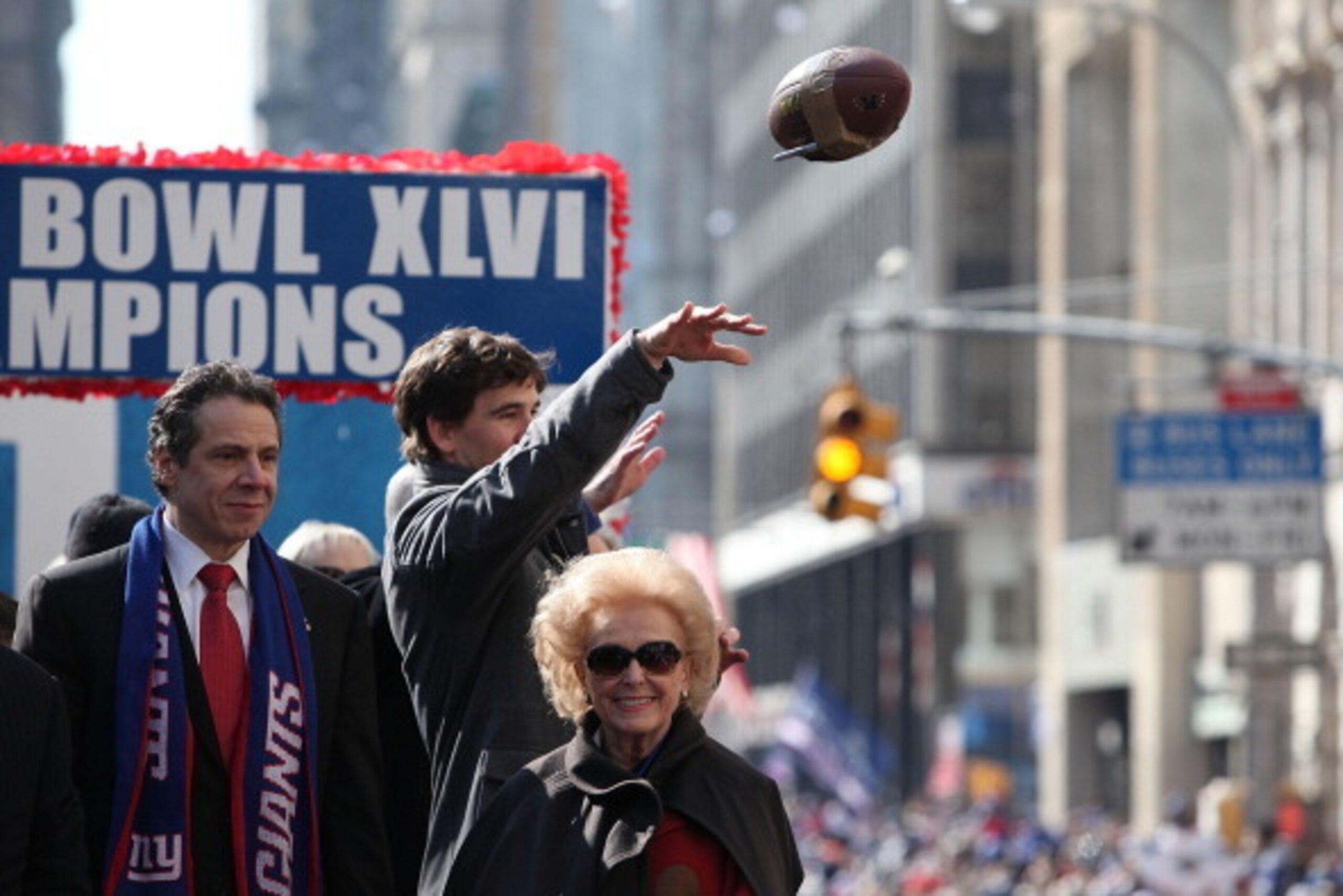 NEW YORK, NY - FEBRUARY 07:  Quarterback Eli Manning #10 (C) of the New York Giants throws back a football that was thrown to him as New York Gov. Andrew Cuomo (L) and Ann Mara looks on during the Giants' Victory Parade on February 7, 2012 in New York Cit