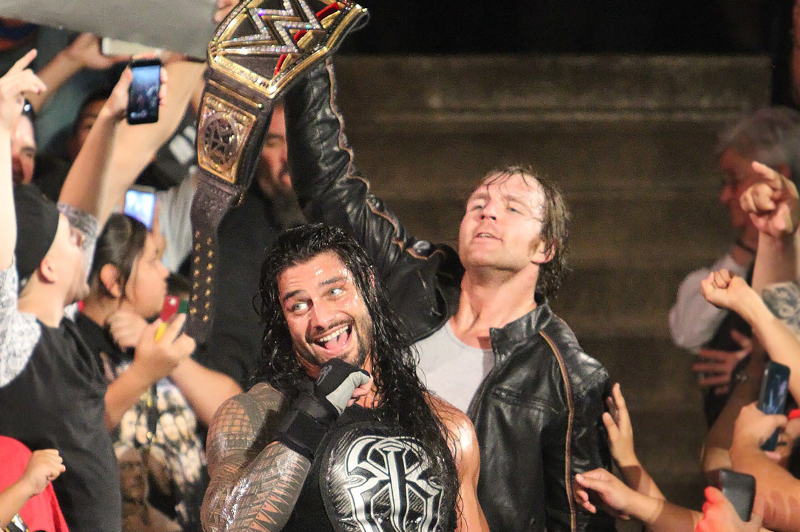 Dean Ambrose holds up the WWE title with Roman Reigns at his side.