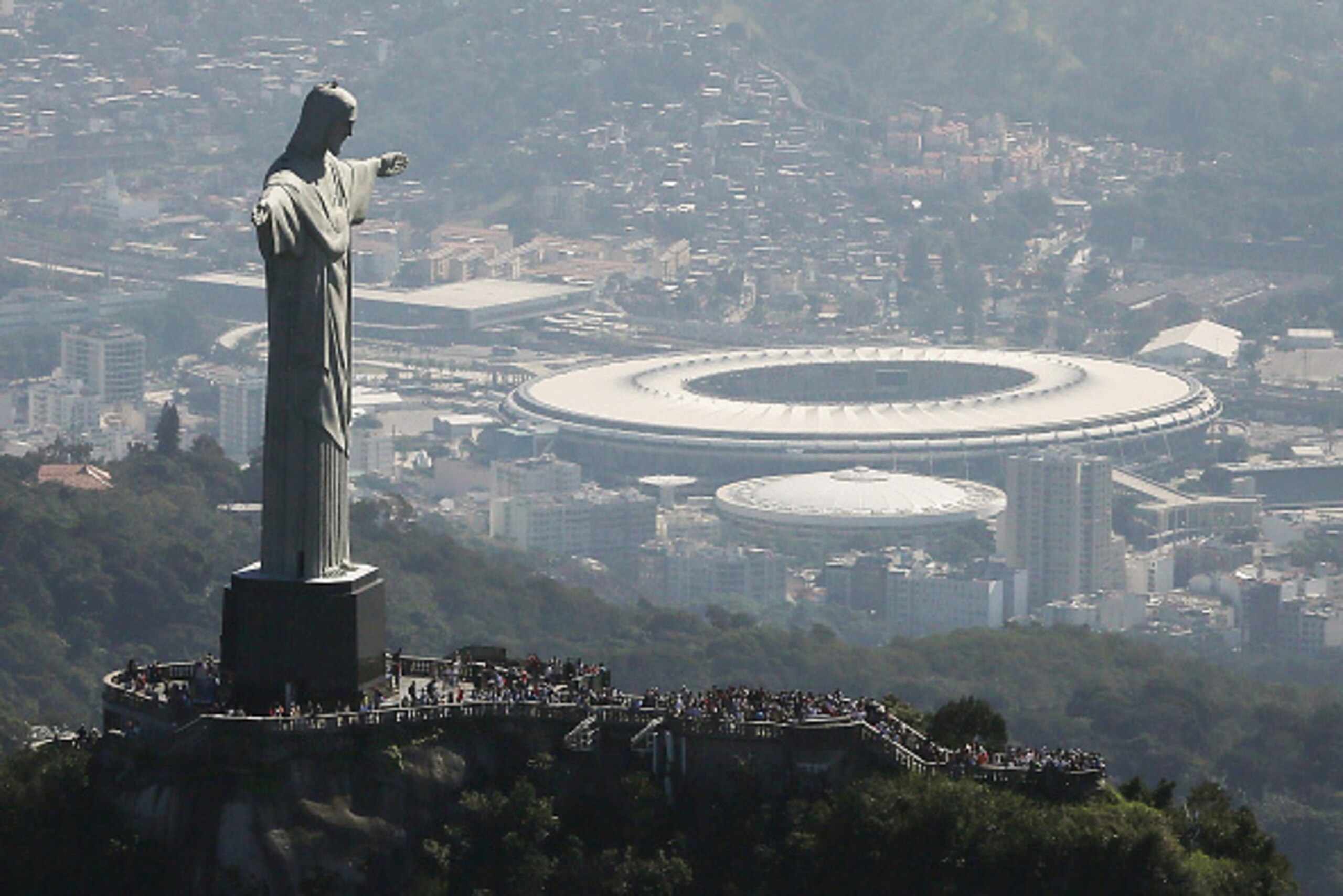 RIO DE JANEIRO, BRAZIL - JULY 04:  Visitors gather beneath the Christ the Redeemer statue as Maracana stadium, site of the Olympic opening ceremonies, stands in the background on July 4, 2016 in Rio de Janeiro, Brazil. July 5 marks the one-month mark to t
