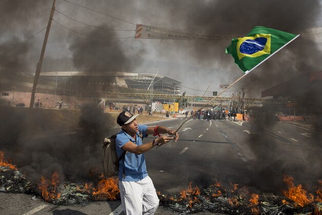 A member of the Homeless Workers Movement waves a Brazilian flag by burning tires during a protest against the money spent on the World Cup near Itaquerao stadium, behind, which will host the international soccer tournament's first match in Sao Paulo, Brazil, Thursday, May 15, 2014. Brazilians are angry at the billions spent to host the World Cup, much of it on 12 ornate football stadiums, one-third of which critics say will see little use after the big event. (AP Photo/Andre Penner)