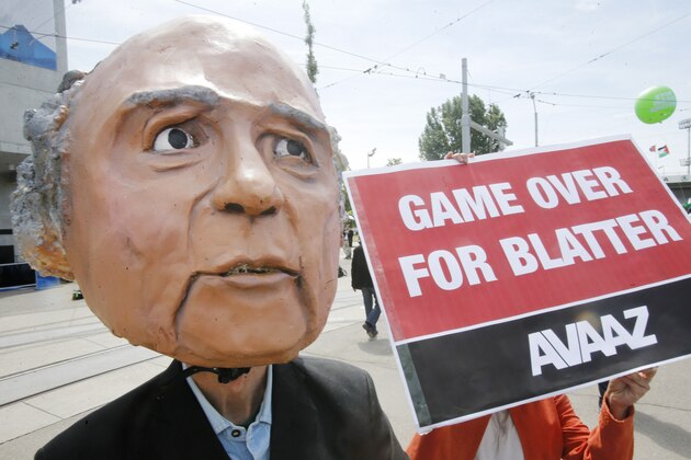 A protester wearing a mask depicting FIFA President Sepp Blatter stands in front of the building where the 65th FIFA congress takes place in Zurich, Switzerland, Friday, May 29, 2015. Protesters from the global campaign movement Avaaz demand the resignation of  Blatter over the FIFA corruption scandal and rights abuses of World Cup construction workers in Qatar. (AP Photo/Michael Probst
