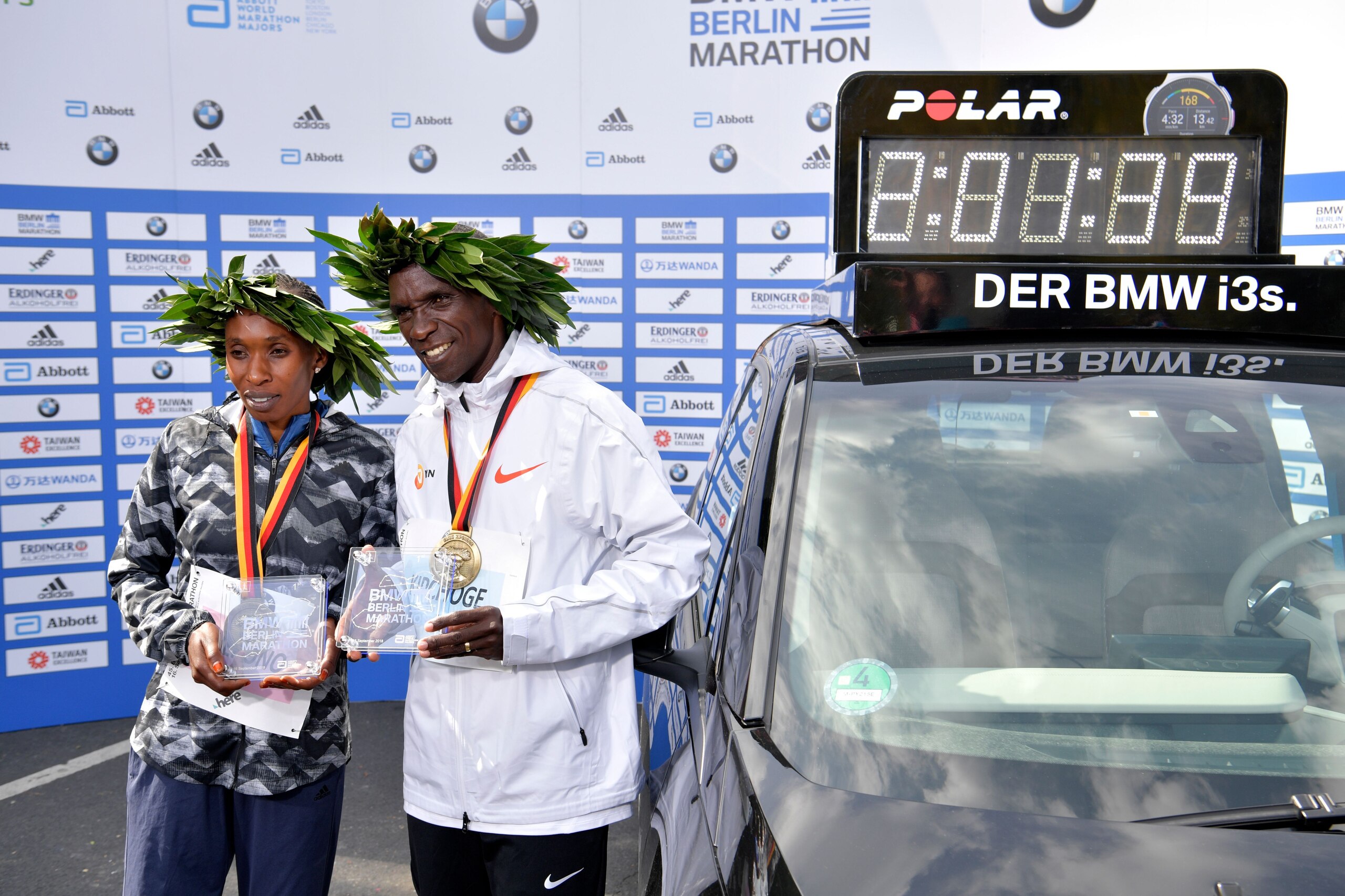 Winner Kenya's Eliud Kipchoge (R) and women's winner Kenya's Gladys Cherono pose with their trophies and medals next to a clock displaying Kipchoge's winning time setting a new record after the Berlin Marathon on September 16, 2018 in Berlin. (Photo by John MACDOUGALL / AFP)        (Photo credit should read JOHN MACDOUGALL/AFP/Getty Images)
