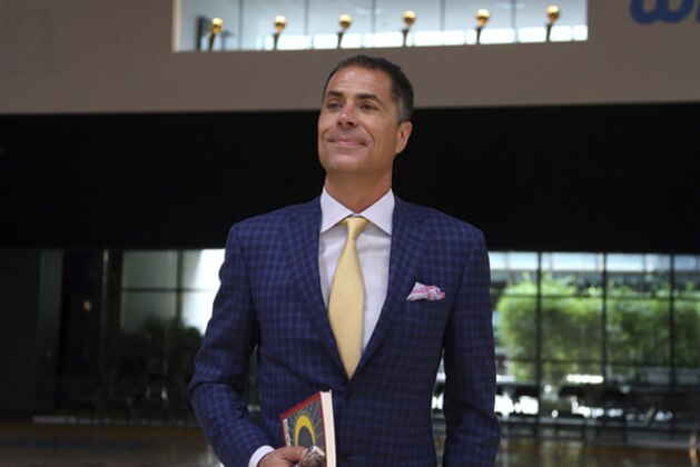 Los Angeles Lakers general manager Rob Pelinka waits to speak about the acquisition of LeBron James and other free agents at a news conference, with Lakers championship trophies visible in a window, above, at the NBA basketball team's headquarters in El Segundo, Calif., Wednesday, July 11, 2018. He's holding a copy of