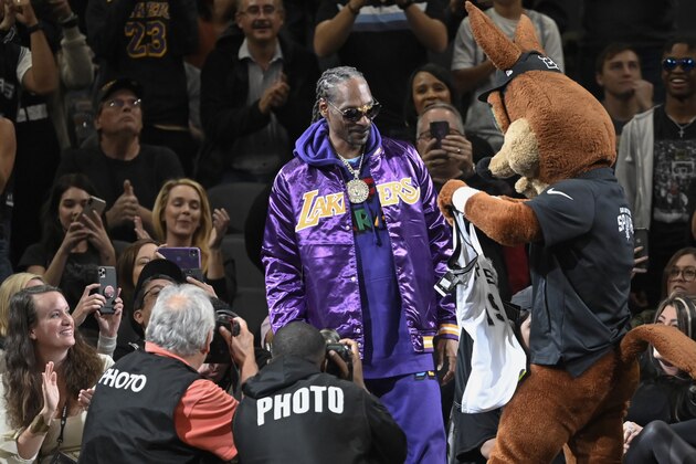 San Antonio Spurs mascot The Coyote, right, presents a team jersey to rapper Snoop Dogg, center, during the first half of an NBA basketball game against the Los Angeles Lakers, Sunday, Nov. 3, 2019, in San Antonio. (AP Photo/Darren Abate)