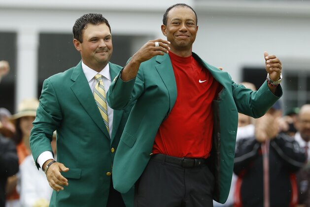 FILE - In this April 14, 2019, file photo, Patrick Reed, left, helps Tiger Woods with his green jacket after Woods won the Masters golf tournament, in Augusta, Ga. The annual rite of spring for golf won't happen this year. The Masters has been postponed until a later date. Augusta National did not indicate when the Masters would be played. That means there will be no golf at least for the next month. The Masters began in 1934 and only World War II has kept it from being played. This was the biggest shoe to drop for golf. The PGA Tour already canceled the next three events leading up to the Masters. Tiger Woods was to be going after his sixth green jacket. (AP Photo/Matt Slocum, File)