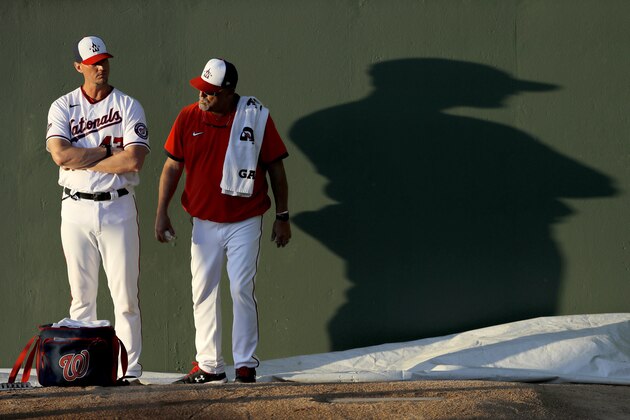 The shadow of Sam Narron, left, pitching coach of the Washington Nationals Double-A Harrisburg Senators, is seen as he talks with Washington Nationals pitching coach Paul Menhart prior to a spring training baseball game against the St. Louis Cardinals, Thursday, March 5, 2020, in West Palm Beach, Fla. (AP Photo/Julio Cortez)
