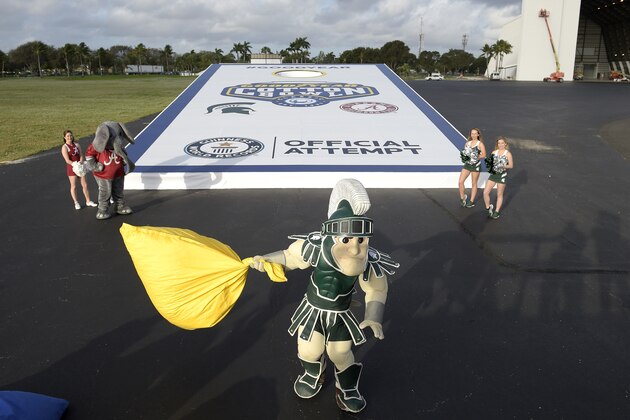 IMAGE DISTRIBUTED FOR THE GOODYEAR TIRE & RUBBER COMPANY - The Michigan State mascot tosses a bean bag, as their cheerleaders and those of Alabama watch, during the World's Largest Game of Cornhole at the Goodyear Blimp Base in Pompano Beach, Fla., Monday, Dec. 28, 2015. The mascots from both universities squared off in advance of the Goodyear Cotton Bowl Classic college football game in the World's Largest Game of Cornhole. The Goodyear Blimp has provided aerial coverage over college football games for the past 60 years and will be providing fans that traditional aerial view over all New Year's Six bowl games in 2015. (Phelan M. Ebenhack/AP Images for The Goodyear Tire & Rubber Company)