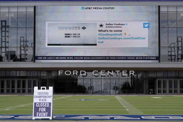 A sign sits on a artificial field frequented by fans at the Ford Center by The Star, the Dallas Cowboys headquarters and training facility, as a large video screen on the exterior wall of the building broadcast the team's most recent news developing from the NFL football draft in Frisco, Texas, Saturday April 25, 2020. (AP Photo/Tony Gutierrez)
