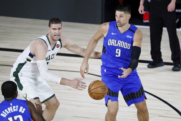 Orlando Magic center Nikola Vucevic (9) passes the ball to forward Gary Clark (12) in front of Milwaukee Bucks center Brook Lopez (11) during the first half of Game 1 of an NBA basketball first-round playoff series, Tuesday, Aug. 18, 2020, in Lake Buena Vista, Fla. (Kim Klement/Pool Photo via AP)